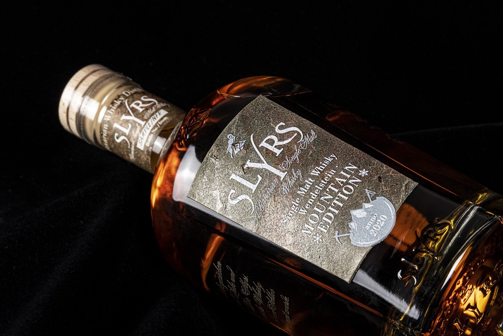 Detailaufnahme einer SLYRS-Flasche mit silbernem Etikett und bernsteinfarbenem Whisky vor schwarzem Hintergrund